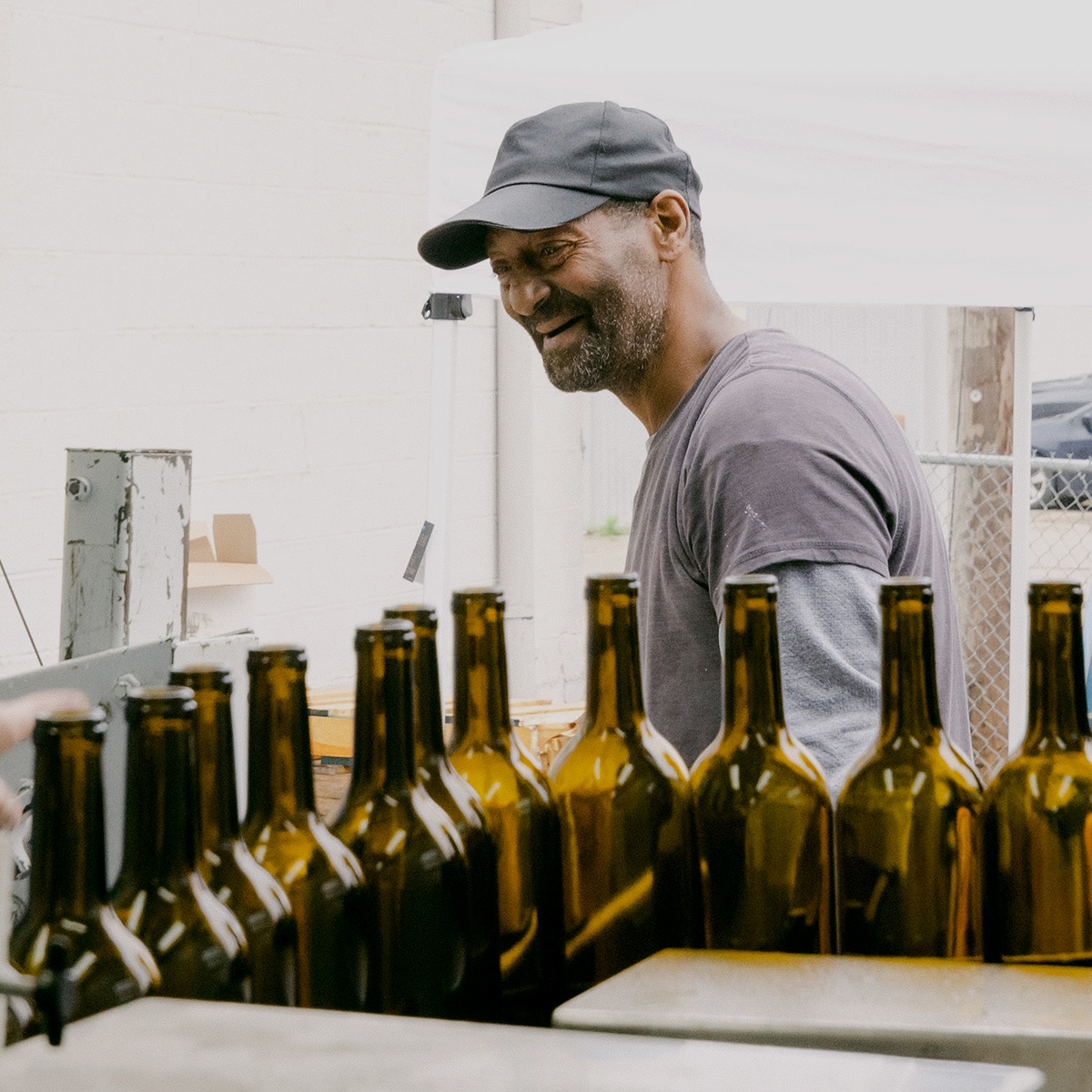 A smiling man in a black cap works at a bottling station with rows of empty wine bottles lined up in front of him.