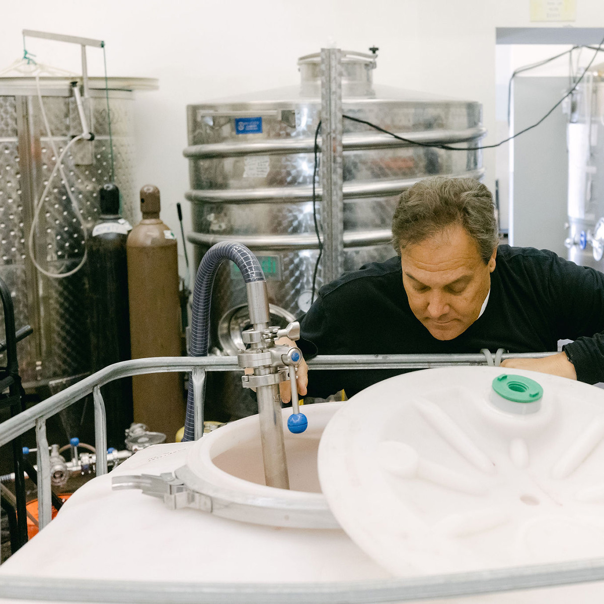 A man carefully monitoring a tank in a winery, surrounded by large stainless steel tanks and industrial equipment.