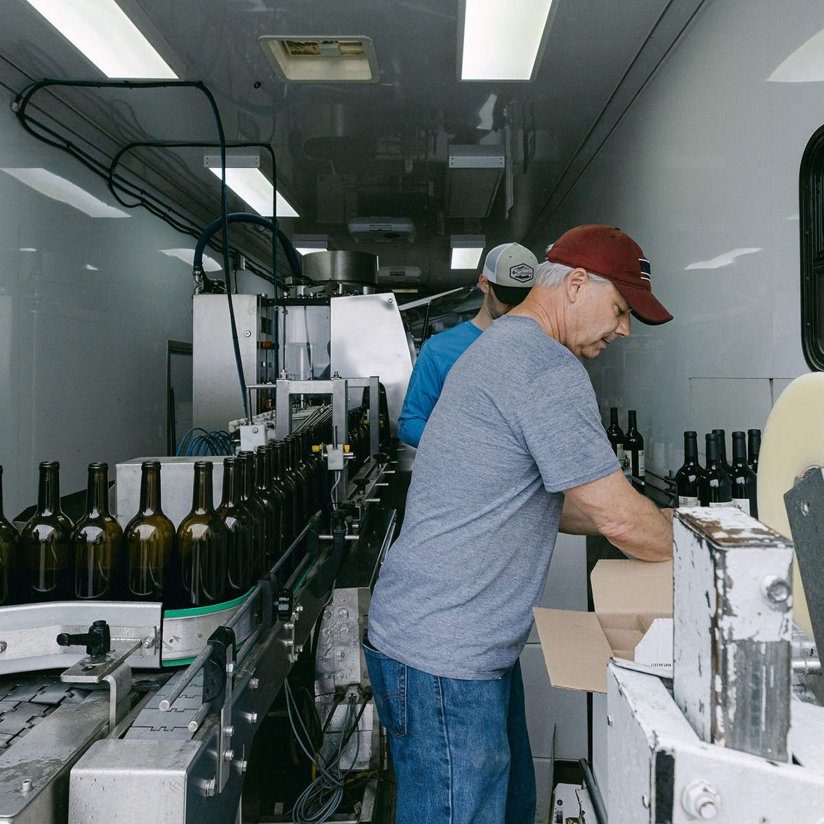 A worker in a red cap and grey shirt operates a bottling line inside a mobile bottling unit, preparing wine bottles for packaging.