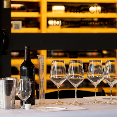 A wine tasting setup on a table with a bottle of red wine, five empty wine glasses, a metal wine chiller, a measuring cylinder, and a pen with paper, set against a backdrop of wine shelves.