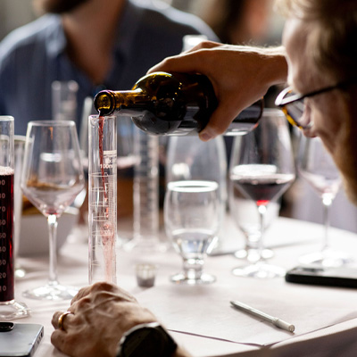 A close-up of a person pouring red wine into a measuring cylinder during a wine blending session.