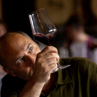 A man carefully examining a glass of red wine, holding it up to the light during a wine tasting event.