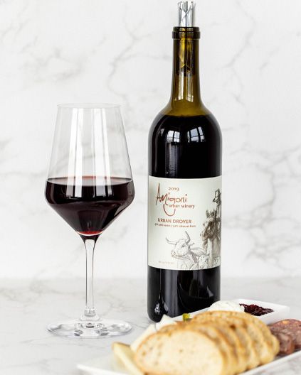 A bottle of Amigoni Urban Winery's 2019 Urban Drover red wine next to a filled wine glass, with a charcuterie board in the foreground on a white marble surface.