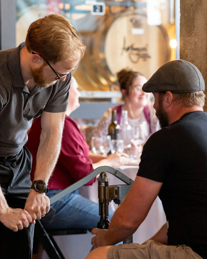 A man with a beard and cap assists another man using a wine bottle corking machine during a hands-on wine event, with other guests seated in the background.