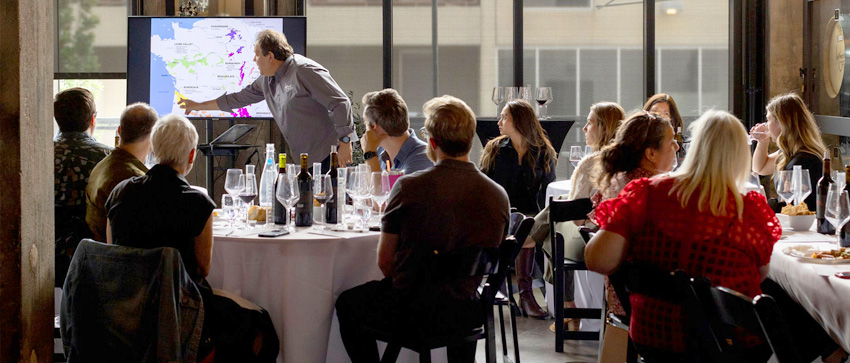 A group of people sitting at round tables in a wine tasting room, listening to a presentation given by a man pointing at a map displayed on a large screen.