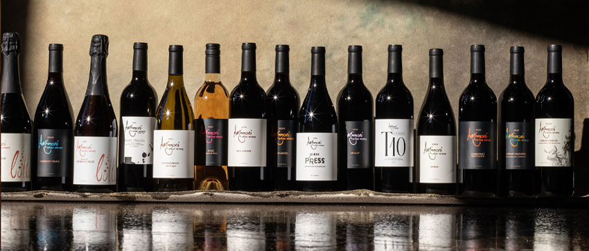 A wide selection of Amigoni Urban Winery wine bottles displayed in a row on a reflective surface with a rustic background.